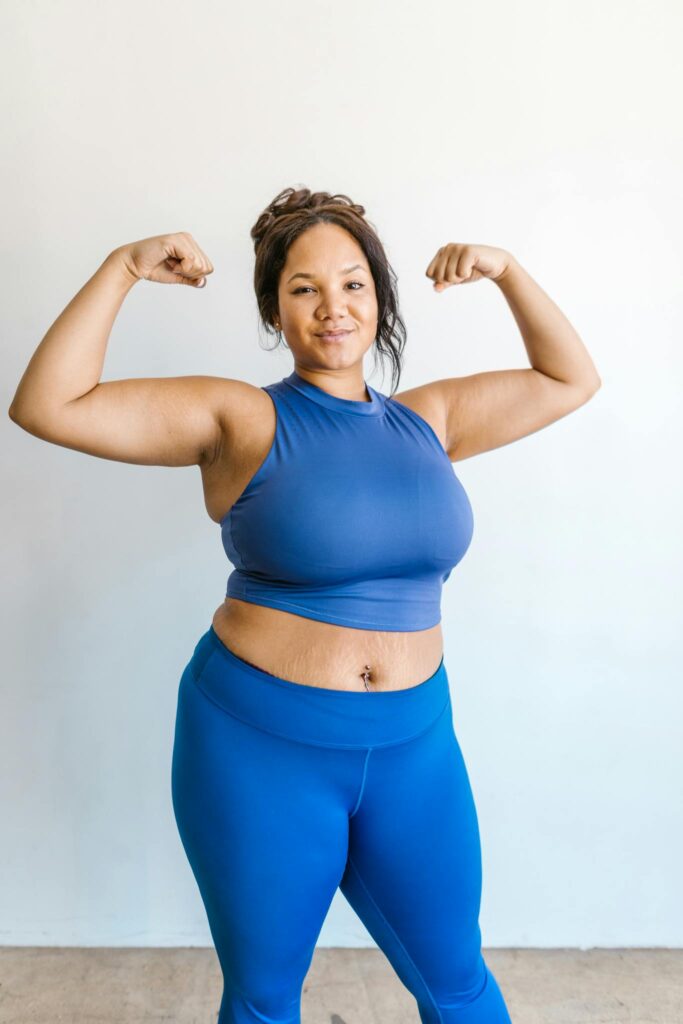 Plus-size woman in blue activewear confidently posing and flexing muscles against a white wall.