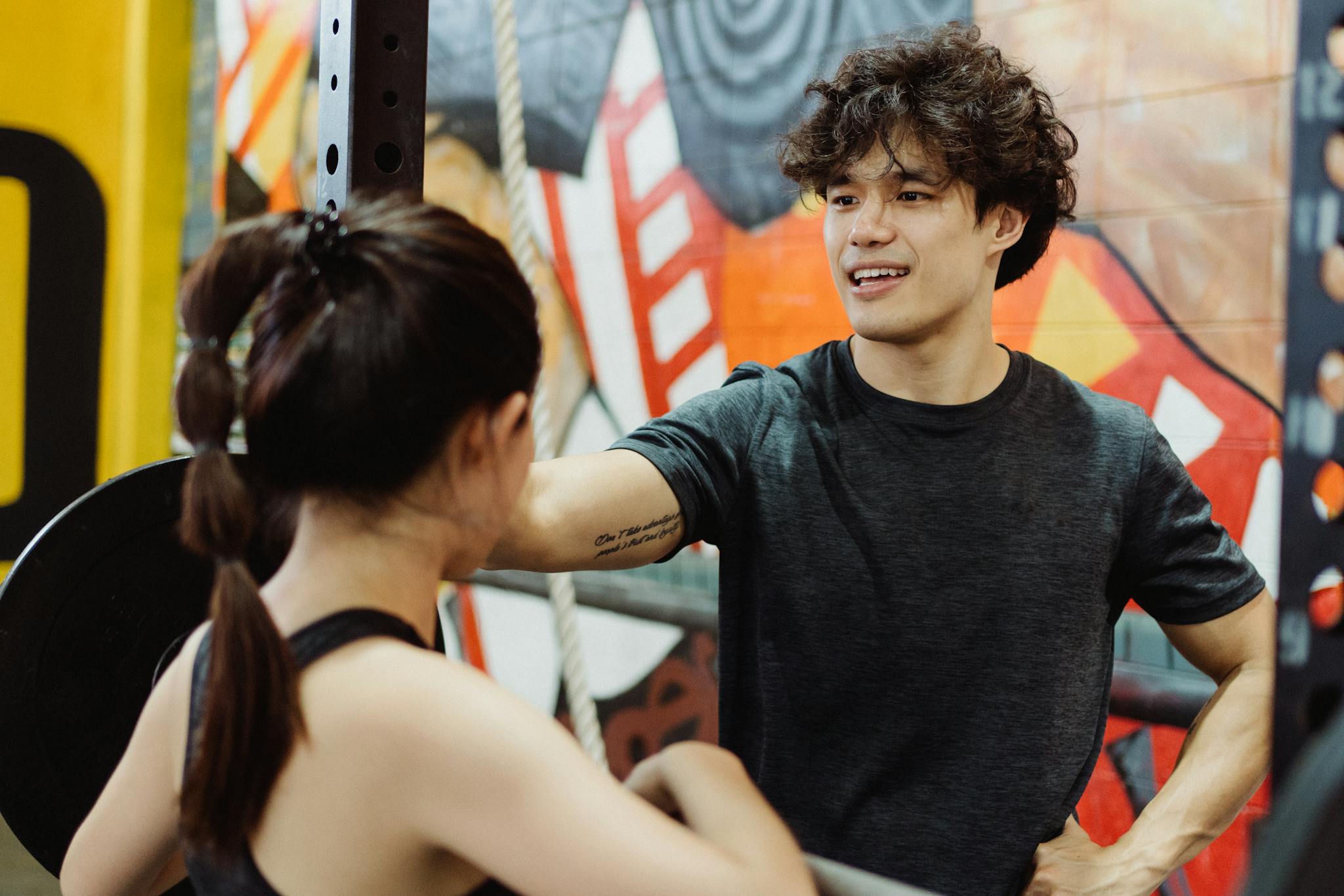 Two young adults in a gym engaging in a lively fitness discussion with a vibrant mural in the background.